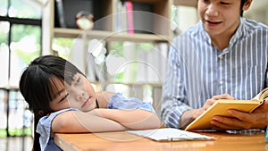 Sleepy little Asian girl leaning on the table while her teacher is teaching and asking a question