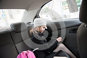 Sleepy little girl in autumn black coat and white hat on trip to school in morning sitting on back seat of safe car with pink