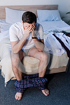 Sleepy man with hands on face sitting on bedding near clothing.