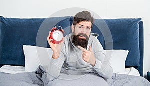 Sleepy man pointing finger at alarm clock being in bed awake, morning