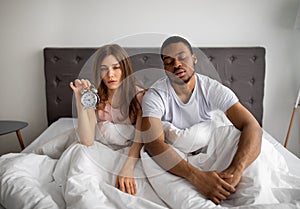 Sleepy multiracial couple sitting on bed with alarm clock, cannot open their eyes, too tired to get up in morning