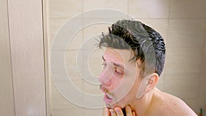 Sleepy shaggy sick young man with flu looks at mirror in bathroom in morning.