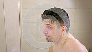Sleepy shaggy tired young man looks at mirror in bathroom in morning.