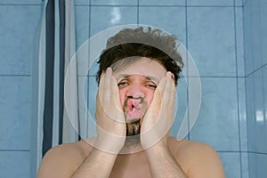 Sleepy shaggy young man with headache in bathroom in morning rubs his face.