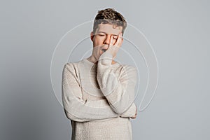 Sleepy teenager boy bored yawning tired covering mouth with hand, standing over gray background. Restless and sleepiness