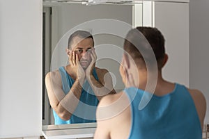 Sleepy tired man in bathroom looks at himself in mirror. Male touching his beard and face. Morning routine