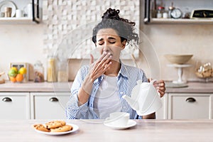 Sleepy tired woman waking up early, pouring tea into cup and yawning, sitting in modern kitchen interior, copy space