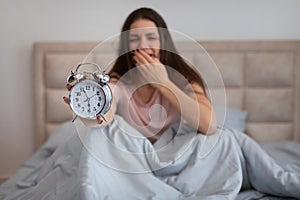 Sleepy woman in bed yawning, holding alarm clock in the morning