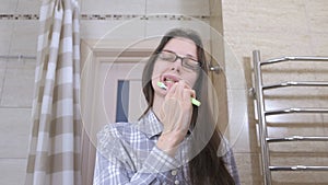 Sleepy woman in glasses in the bathroom cleaning her teeth with toothbrush.