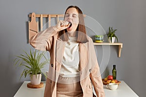 Sleepy worker. Early awaking. Yawning woman with brown hair wearing beige jacket standing in home kitchen interior covering mouth