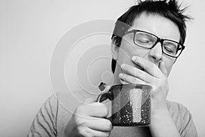 Sleepy yawning man in eyeglasses with red cup of tea or coffee has uncombed hair in underwear on light background, morning refresh