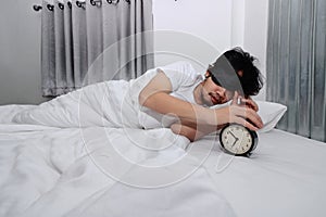 Sleepy young Asian man with eye mask touching turn it off to alarm clock in comfort bed