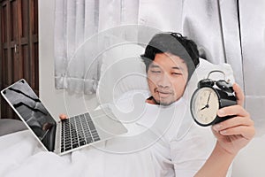 Sleepy young Asian man turns off the alarm clock on the white bed in bedroom. Working hard to meet deadlines concept