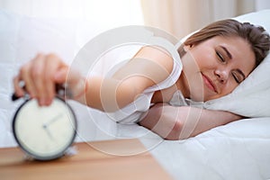 Sleepy young brunette woman stretching hand to ringing alarm willing turn it off. Early wake up, not getting enough