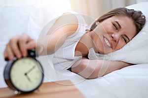 Sleepy young brunette woman stretching hand to ringing alarm willing turn it off. Early wake up, not getting enough