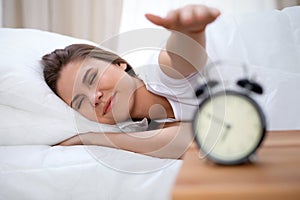 Sleepy young brunette woman stretching hand to ringing alarm willing turn it off. Early wake up, not getting enough