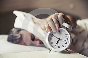 Sleepy young man covering ears with pillow as he looks at alarm clock