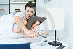 sleepy young man reaching for alarm clock while sleeping with girlfriend