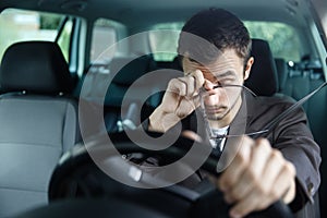 Sleepy young man rubs his eyes with his right hand. His left hand is on the steering wheel. He is sitting at his car. Road safety photo