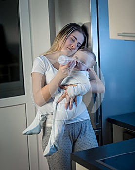 Portrait of sleepy young mother feeding her baby boy with milk from bottle at night