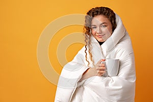 Sleepy young woman with curly hair covered in blanket and holding cup of coffee against yellow background
