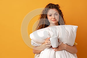 Sleepy young woman with curly hair covered in blanket and holding cup of coffee against yellow background