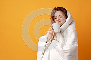 Sleepy young woman with curly hair covered in blanket and holding cup of coffee against yellow background