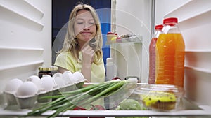 Sleepy young woman opens the refrigerator door at night, looks inside and picks out orange juice. View from inside the