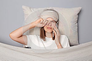 Sleepy young woman wearing white T-shirt and sleep eye mask lies under blanket in bedroom isolated on gray background talking on