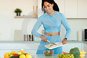 Slender african american woman in sportswear standing in the kitchen and preparing fresh vegetable salad