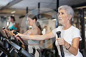 Slender athletic mature woman during cardio exercises on elliptical simulator in fitness center