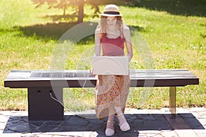 Slender attrective girl looking for information in net, laptop charging with help of solar panel built in wooden bench, using