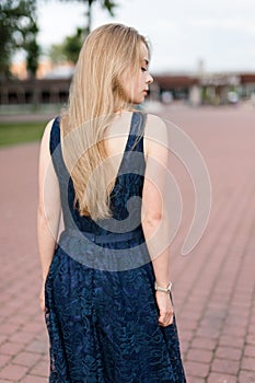 Slender blonde girl in dark-blue dress turned her back looking away on pavement