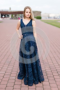 Slender blonde girl in elegant dark-blue floor-length dress on the pavement