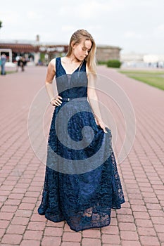 Slender blonde girl touching hem of elegant dark-blue floor-length dress on the pavement