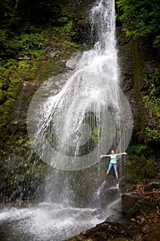 A slender blonde stands under a large picturesque waterfall, spreading her arms to the sides
