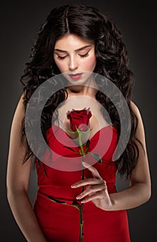 A slender brunette woman with long curly hair and beautiful makeup, she is wearing a red dress, she is holding a rose