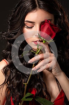 A slender brunette woman with long curly hair and beautiful makeup, she is wearing a red dress, she is holding a rose