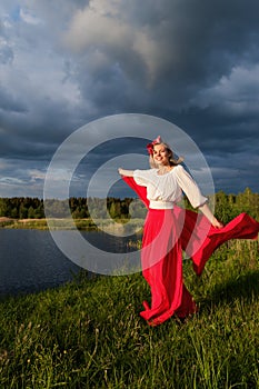 A slender caucasian girl in red skirt and white blouse with a red shawl in her hands against the background of a stormy sky