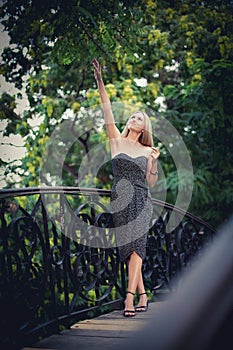 A slender elegant girl in a dark dress with blond hair stands in a park alley on a bridge