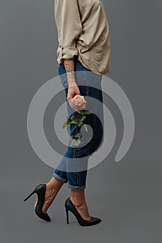 A slender girl in classic black high-heeled shoes and jeans holds in her hand with a pink rose tattoo around a dark