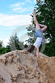 slender girl does handstand in open air against background of blue sky. Female athlete in white T-shirt and denim shorts performs