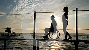 Slender girl with man disabled on wheelchair walking on pier at sea against sky in sunset