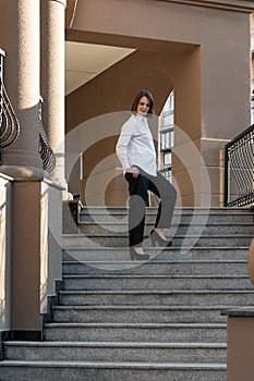 Slender girl is photographed on steps of beautiful building. Young woman is dressed in classic white shirt and black pants