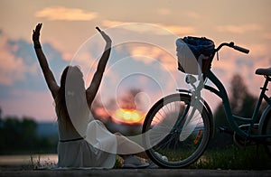 Slender girl sits with her back arms raised near retro bicycle with basket enjoying colorful sky with clouds at sunset