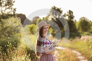A slender girl in a straw hat and with a straw handbag holding a bouquet of daisies in the meadow. Summer sunset, the concept of