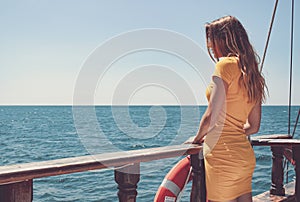 A slender girl in a yellow dress looks out over the horizon from an vintage historical ship.