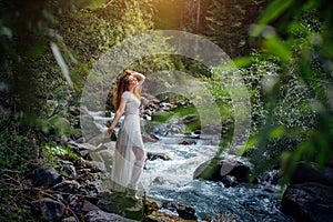 Slender long-haired brunette in white dress posing against small mountain river and green trees. Beautiful young woman walking