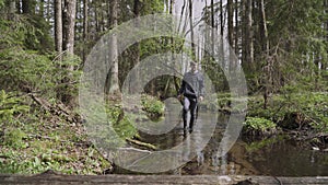 Slender man in boots carefully walks along bed of clean river in wild forest.