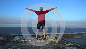 A slender man in a red jacket stands on a high mountain in a strong wind and depicts a flight with outstretched arms